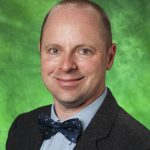 Color photograph of Prof. Clark A. Pomerleau, a clean-shaven white man in his forties with short-cropped brown hair and smiling hazel eyes. He is dressed in a light blue button-down shirt, navy bowtie, and tweedy grey suit jacket. The photo's background is green.