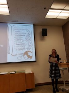 photo of Elizabeth Donaldson speaking adjacently to a projected image of a panel from Ellen Forney's graphic memoir on bipolar disorder, “Marbles.” Elizabeth is wearing a black and white ensemble that echoes the black and white ink drawing depicted in the book.