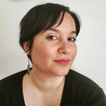 Color photograph of Prof. Shayda Kafai, a Middle Eastern femme wearing bright pink lipstick. She smiles at the camera. She has short, wispy hair, a long silver feather earring, and is wearing a black shirt.